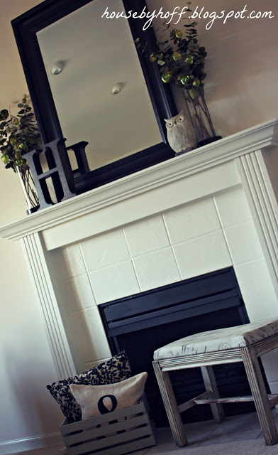The same fireplace but painted a white color with the mantel painted as well. A black mirror is on the mantel and a foot stool is in front of the fireplace.