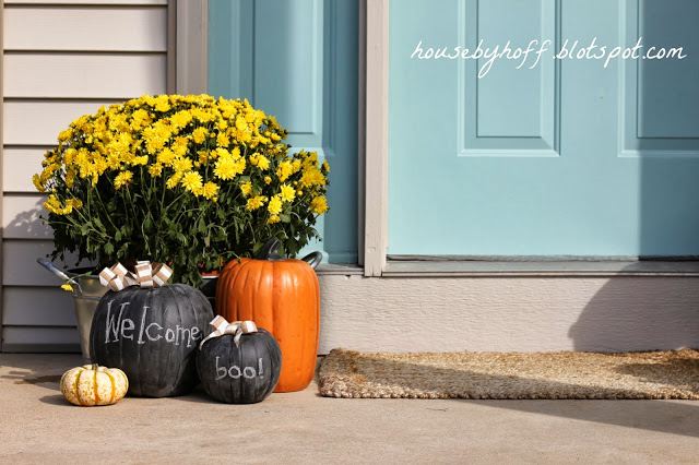 Front porch with light blue door, yellow flowers in a planter, and fake pumpkins.