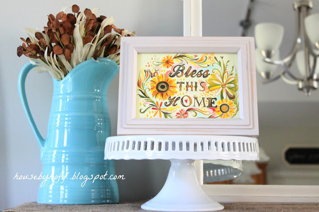 Bless this home sign on a white cake stand on the mantel.