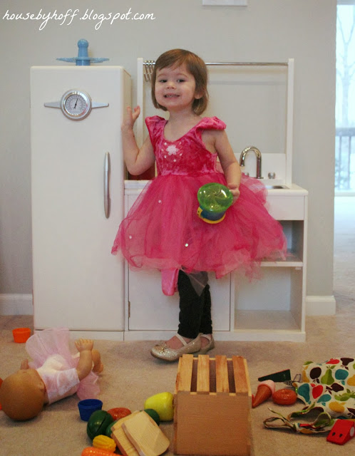 A little girl in a pink tutu dress waving in her playroom.