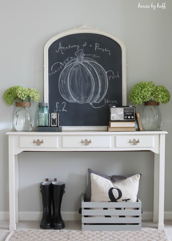 Chalkboard on side table with faux green flowers in clear vases.