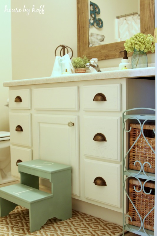 White bathroom cabinets with light green stool and wooden mirror.