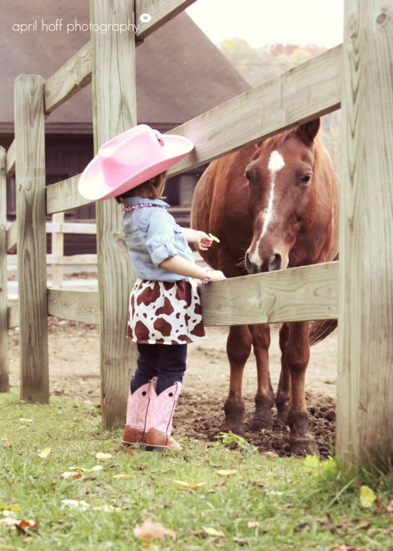 Girl petting a horse in her cowboy boots by a fence.