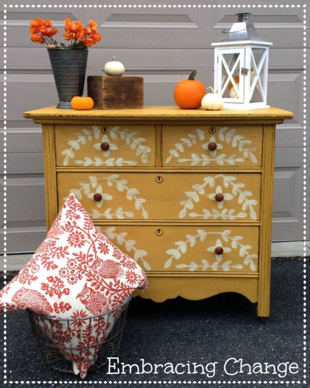 A chippy yellow dresser with pumpkins and a lantern on top of it.