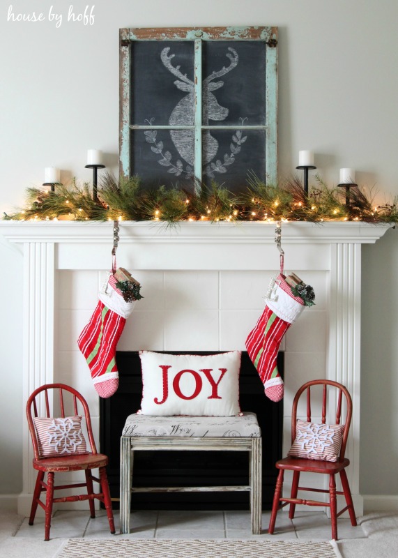 A white fireplace with two stockings hanging and two chairs in front of it, there a chalkboard with a deer drawn on it on the mantel.