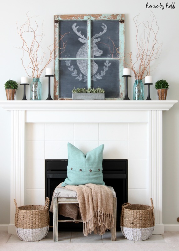 A white fireplace with a bench in front of it, a blue pillow on the bench and a chalkboard deer on top of the mantel.