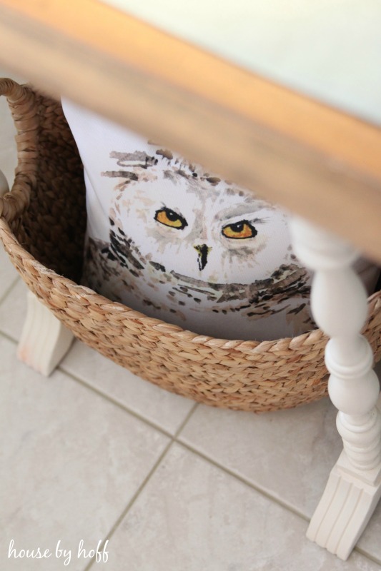 A wicker basket with an owl pillow inside it under the table.