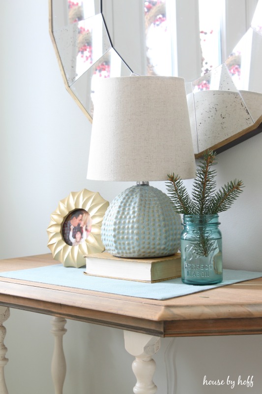 A side table with a small blue lamp and a small vase with evergreen inside it.
