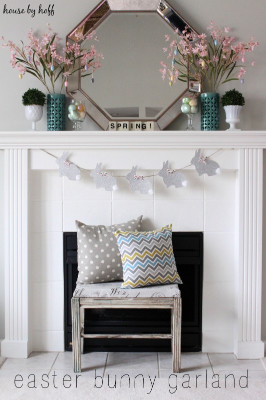 A white fireplace with a mirror on it flanked by vases of pink flowers and a bunny banner in front.