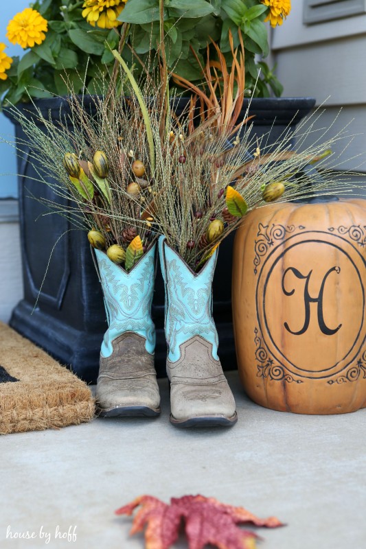 Cowboy boots filled with stalks of decorative grass, a pumpkin, and yellow flowers on front porch.