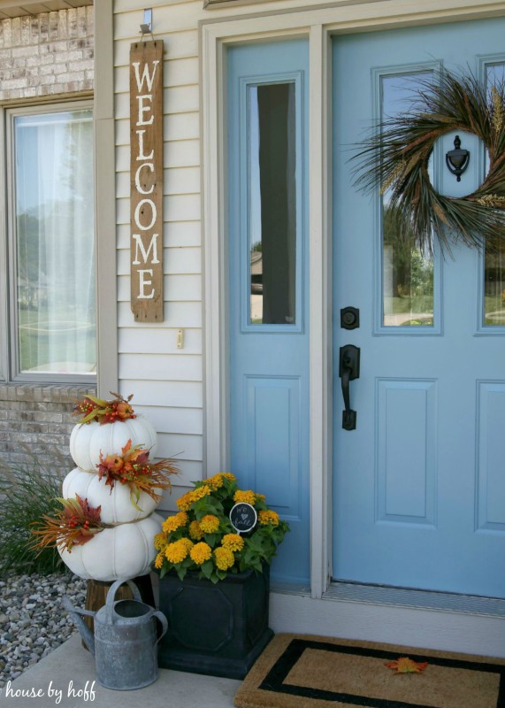 White stacked pumpkins, yellow flowers and a watering can on the front stoop.