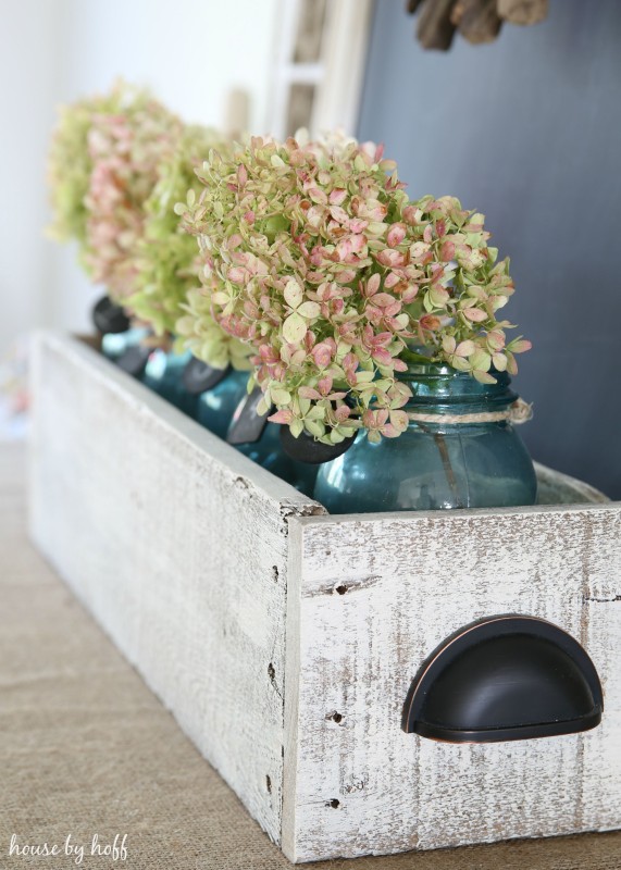 Four glass blue jars in a wooden box filled with hydrangeas.