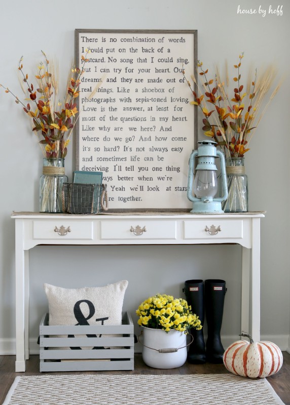 A white pot of yellow flowers under the side table, fall leaves, and rubber boots under the side table.