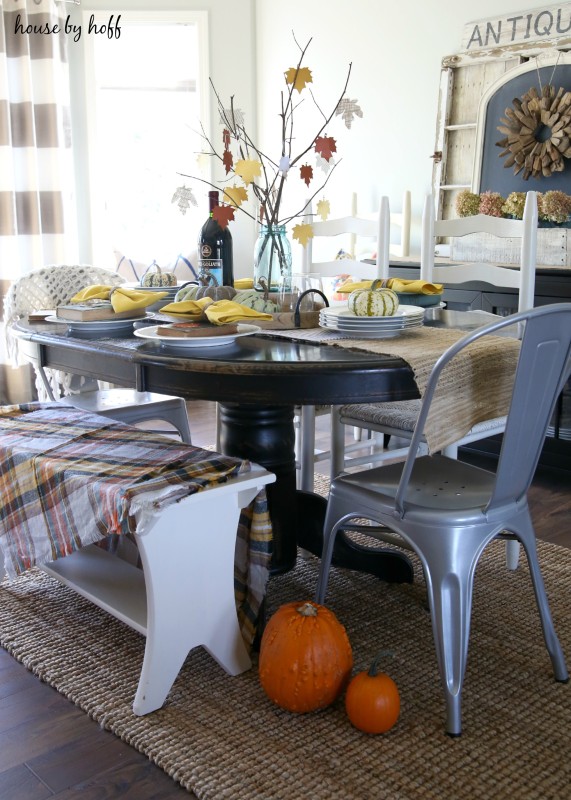 The dining room table, with a bench and a plaid blanket on it, plus two pumpkins on the floor beside the table.