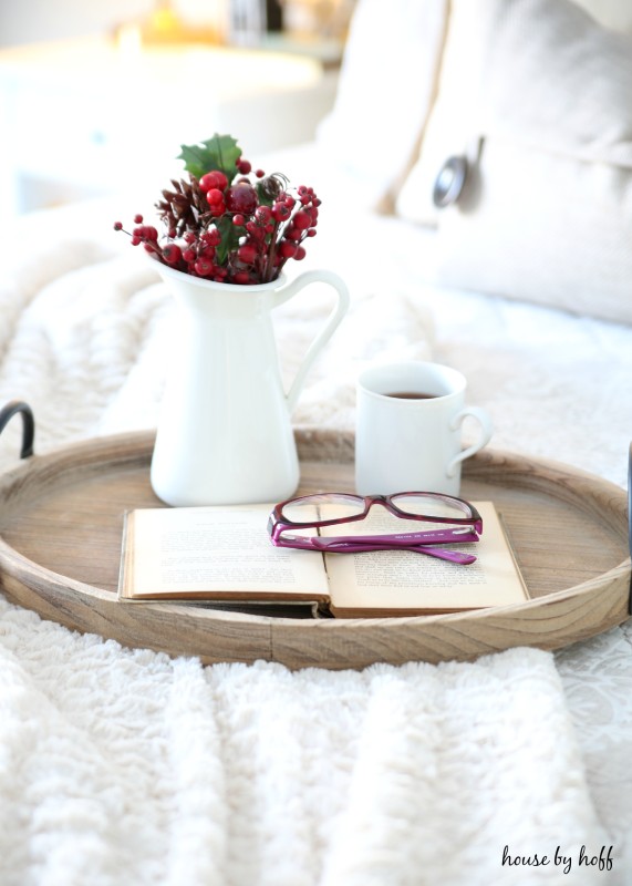 A cut of tea, berries, glasses and a book on the wooden tray on the bed.