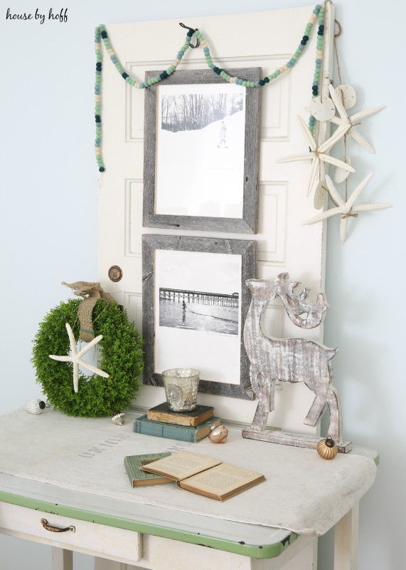 Wooden desk with a green Christmas wreath and starfish on it, a wooden deer, land pictures in wooden frames.