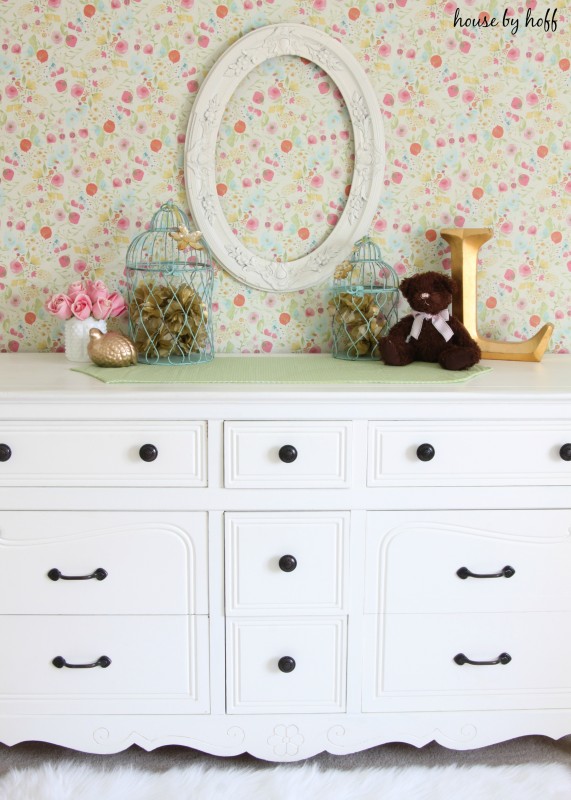 White dresser, teddy bear and pink flowers in front of the wall paper.