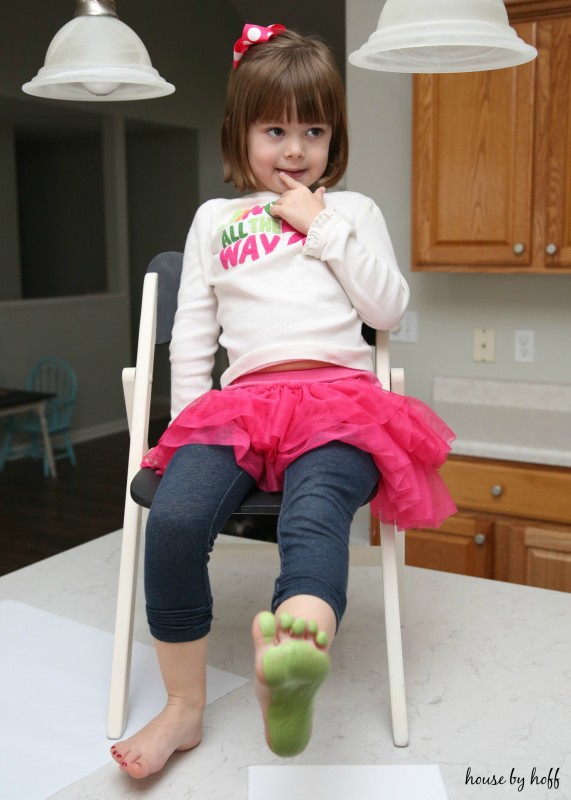 A little girl sitting on a chair with a green foot painted about to put it on the paper.