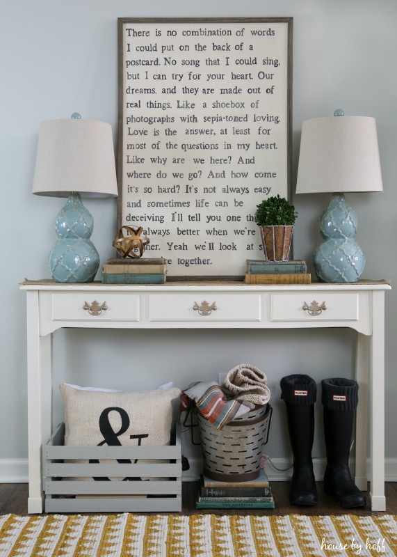 A hallway table with boots, pillows and blankets underneath it and a spoken word poster on top of the table.