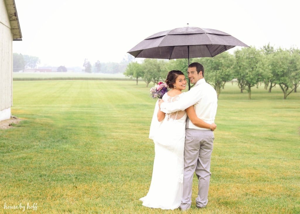 Bride and groom in a field with rain and an umbrella.