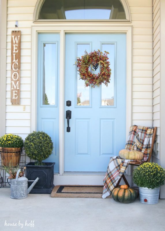 Front porch of house with a light blue door, chair, pumpkins and flowers.