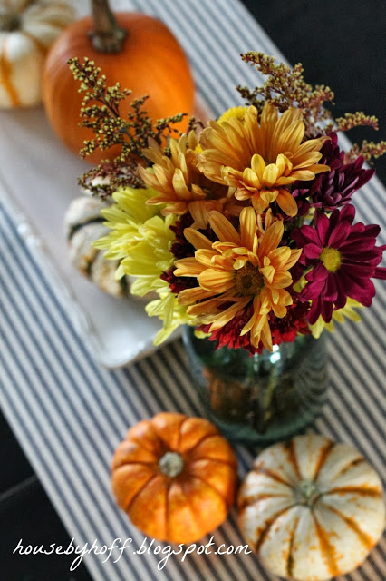 Orange, red, rust and yellow flowers in vase with pumpkins around it on table.