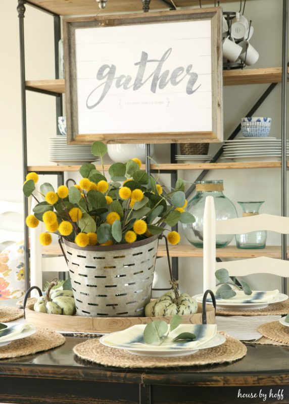 Bright yellow flowers in a steel bucket on table with a gather sign hanging up behind table.