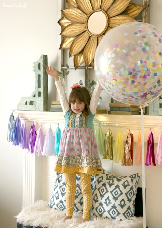 Little girl standing on a bench in front of the fireplace mantel with her arms in the air.