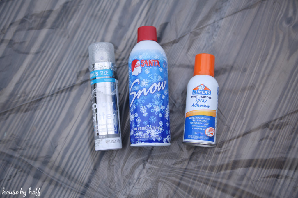 A bottle of spray adhesive, a bottle of snow and a bottle of spray glitter lying on the porch outside.