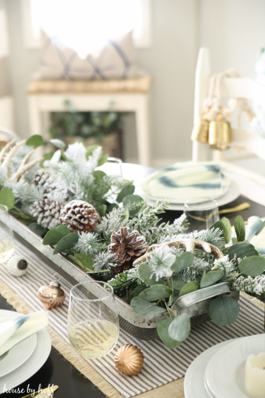  Table set for Christmas with a natural greenery and pine cone centrepiece, Christmas ornaments on the table and white plates.