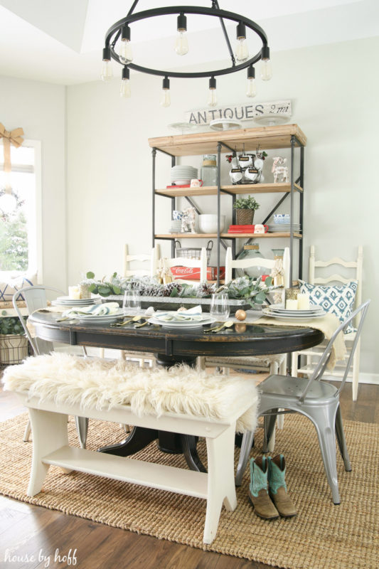 Winter wonderland tablescape with a wooden table with a white bench with faux white fur on it, cowboy boots and an open shelf in dining room.