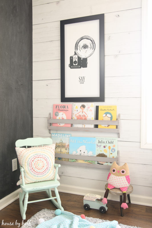 A small blue rocking chair with a pillow and a cute stuffed animal, also books on a shelf on the wall.