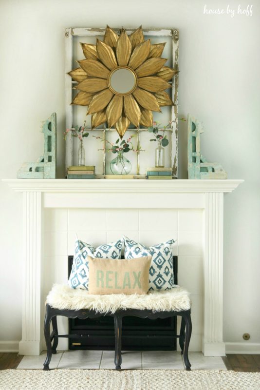 A starburst mirror on the fireplace mantel with a small bench in front of the fireplace and pillows on the bench.