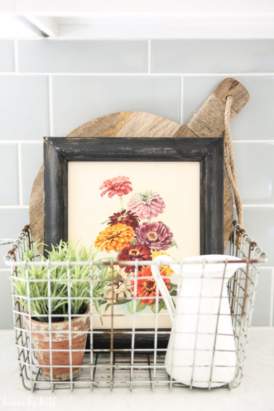 A small floral picture in a wire basket on the counter in the kitchen.