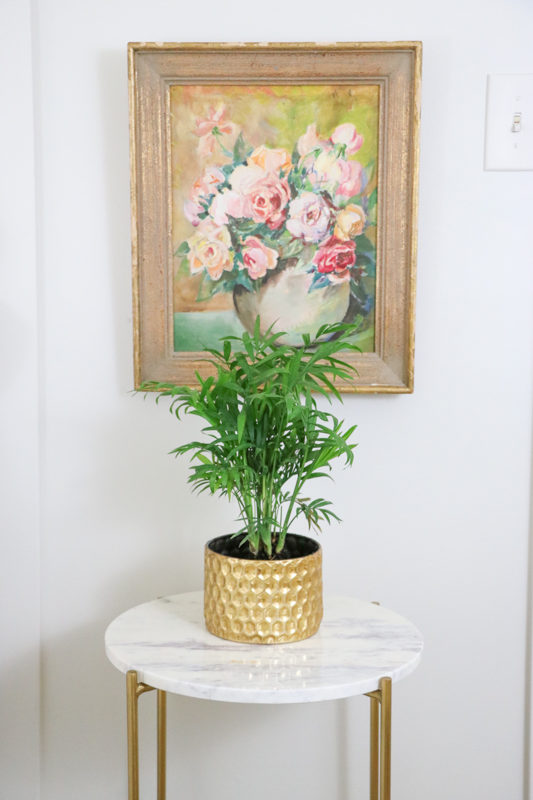 Vintage floral piece hanging just above a side table with a green plant on it.