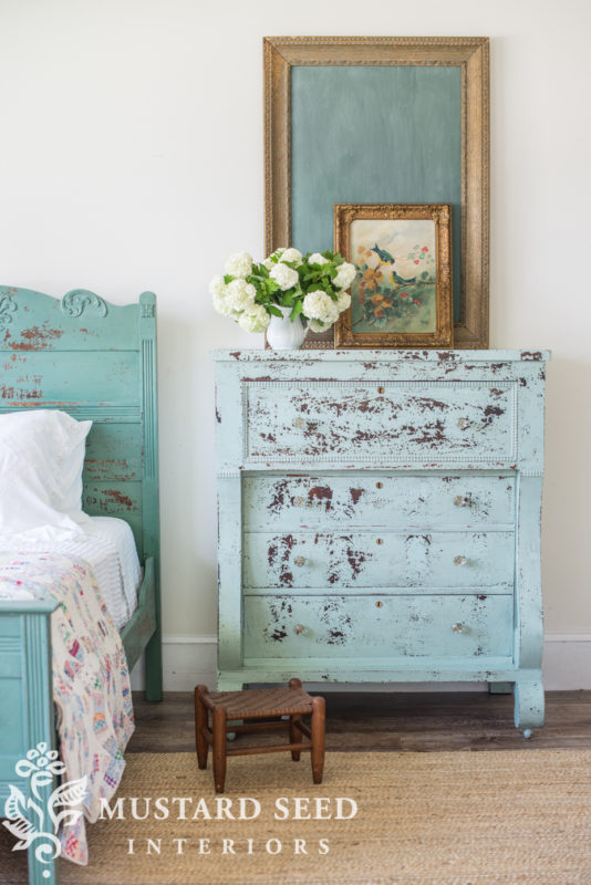 A distressed dresser in the bedroom with a small flower inspired art piece on top.