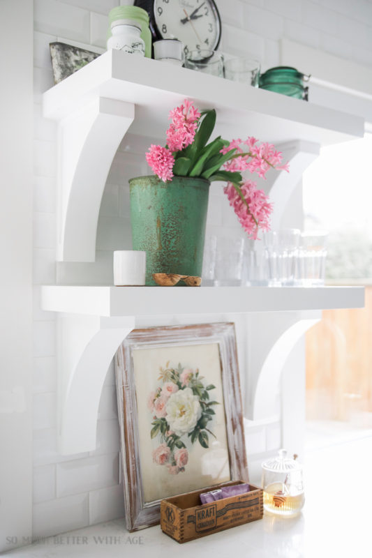 Open shelving in a white kitchen with a vintage looking floral picture on the counter.