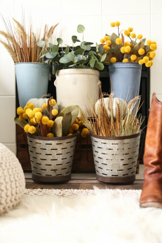 Multi colored buckets full of fall flowers and leaves in front of fireplace.