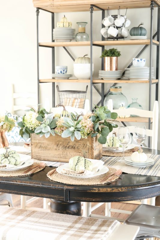 An old wooden crate with white and green hydrangeas in it on a table with plates and pumpkins.