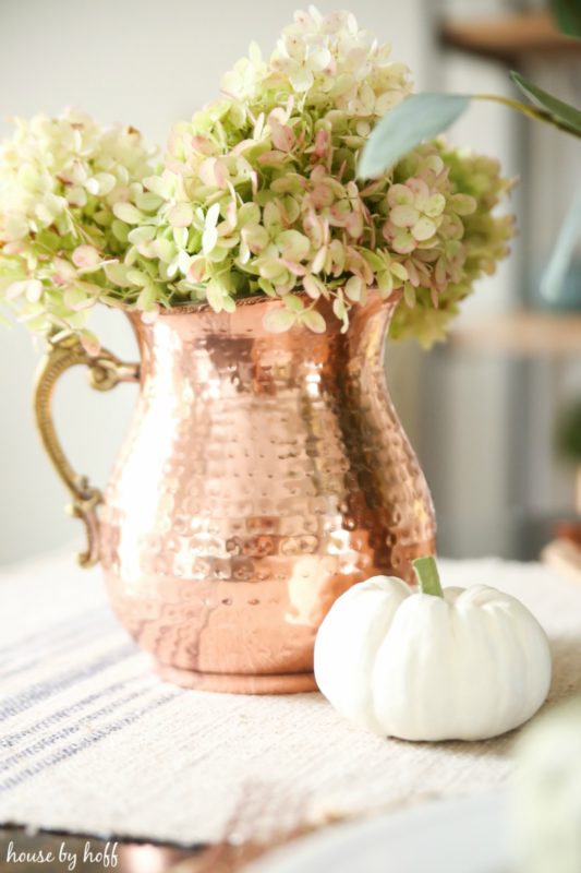 Large copper pitcher with hydrangeas in it and a small white pumpkin beside it.