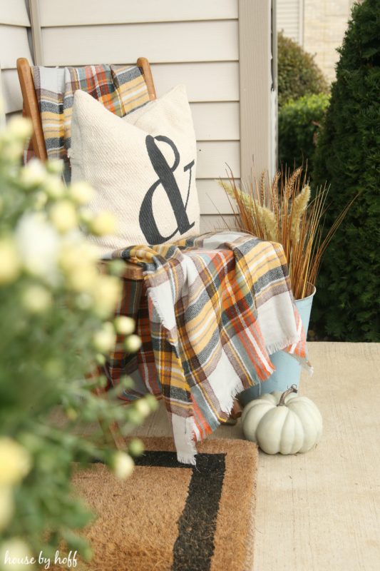 Front porch wooden chair with a tartan blanket and white pumpkin in front of it.