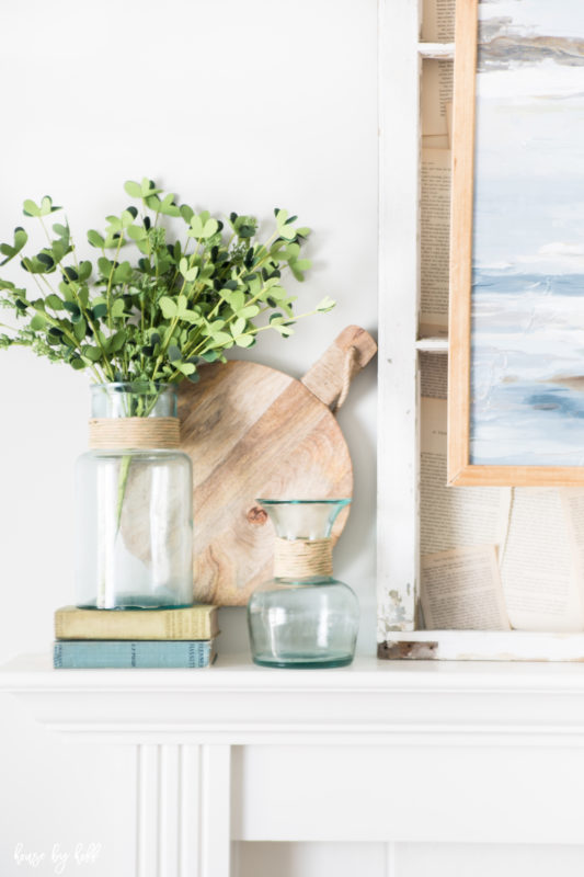 Clear blue glass with branches of greenery in it, sitting on antique looking books on the mantel.