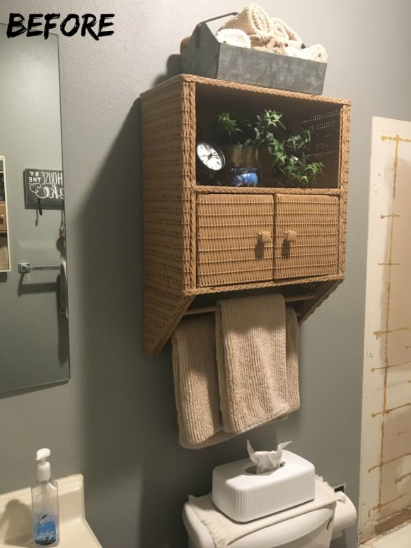 A picture of the bathroom before with dark gray walls and a wicker cabinet on the wall.