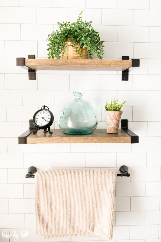 Three small wooden shelves with a clock, a plant, glass and a towel on them in the bathroom.