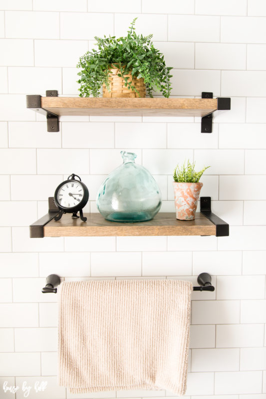 Wooden shelves on wall with a plant, clear glass and small hand towel on them.