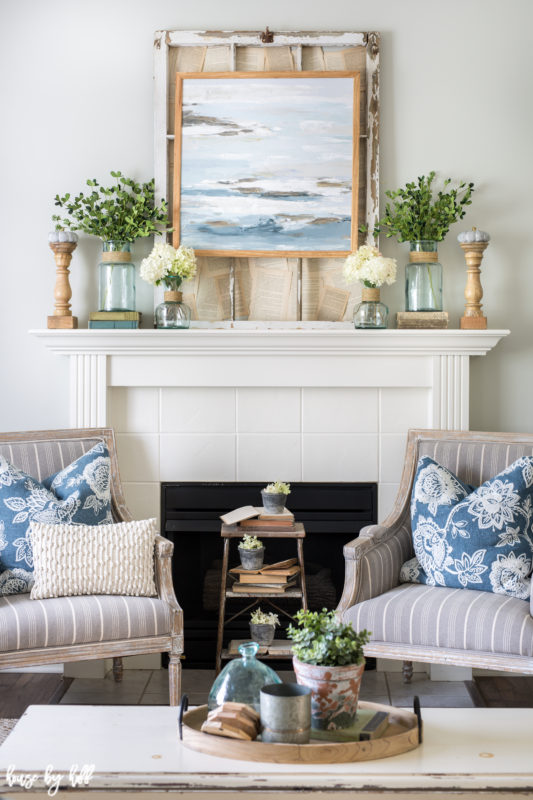 White fireplace with a ocean picture above it and 2 armchairs beside the fireplace.