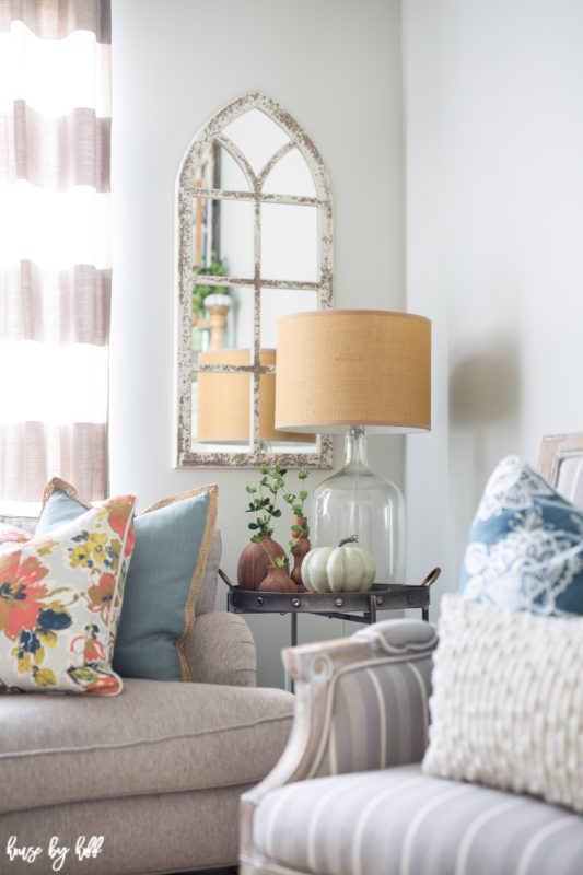 Little white and brown pumpkins on a side table with a lamp on it and a mirror on the wall.