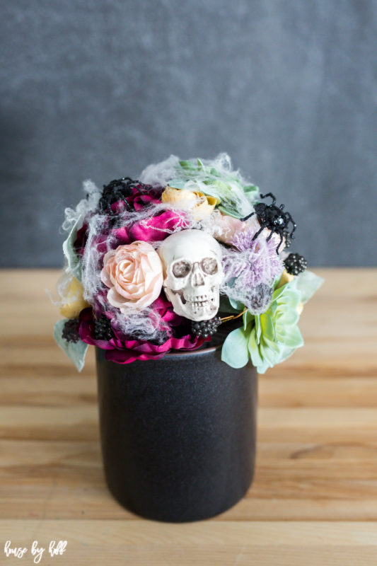 Succulents and flowers with spider web around them and skulls all in a black vase on a table.