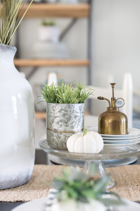 Brushed brass olive oil canister on a white plate with succulents in a silver container on the table.