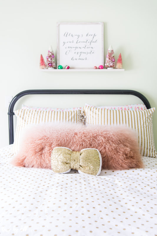 Little bed with fluffy pink pillow and gold bow in room.